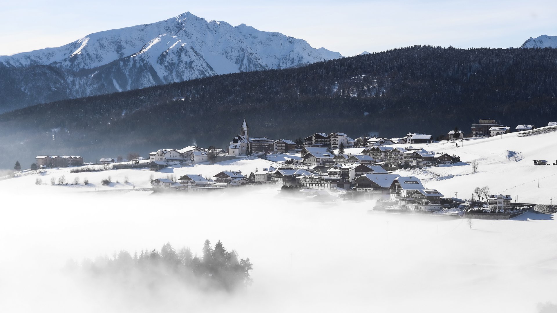 Conoscete già Maranza? L'immagine mostra un villaggio innevato in una valle, circondato da una fitta nebbia. Sullo sfondo si vedono alte montagne innevate e un pendio boscoso. Una chiesa spicca al centro del villaggio e il cielo è sereno e azzurro.