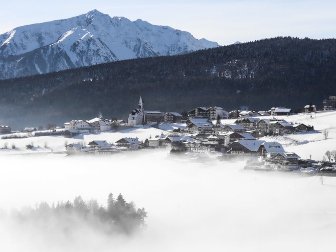 Le località dell’area vacanze Rio Pusteria L'immagine mostra un villaggio innevato in una valle, circondato da una fitta nebbia. Sullo sfondo si vedono alte montagne innevate e un pendio boscoso. Una chiesa spicca al centro del villaggio e il cielo è sereno e azzurro.