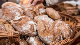 Shopping a Rio di Pusteria (BZ) L'immagine mostra un cesto pieno di pani appena sfornati con una crosta croccante e una superficie leggermente infarinata. Una mano sta prendendo uno dei pani, il che indica una vendita o una selezione.
