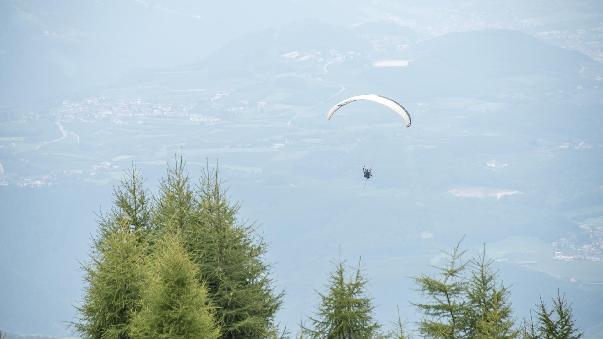 Parapendio in Alto Adige L'immagine mostra un parapendio che vola alto nel cielo sopra una montagna boschiva. Sullo sfondo si vedono dolci colline e una valle con villaggi sparsi e campi.