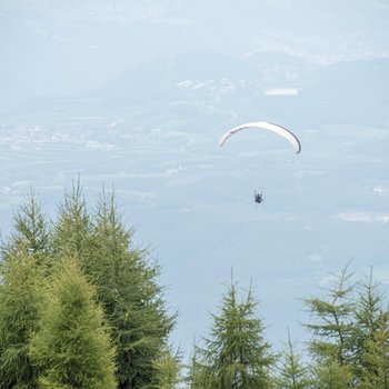 Benvenuti nell’Area Vacanze Sci e Malghe Rio Pusteria! L'immagine mostra un parapendio che vola alto nel cielo sopra una montagna boschiva. Sullo sfondo si vedono dolci colline e una valle con villaggi sparsi e campi.