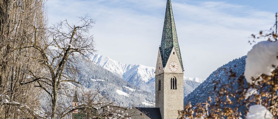 Le località dell’area vacanze Rio Pusteria L'immagine mostra una chiesa con un alto campanile verde in un villaggio innevato. Sullo sfondo si vedono montagne coperte di neve e un cielo azzurro, mentre in primo piano ci sono alberi spogli e cespugli coperti di neve.