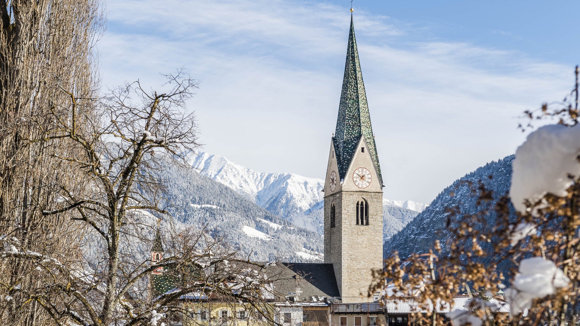 Alla scoperta di Rio di Pusteria L'immagine mostra una chiesa con un alto campanile verde in un villaggio innevato. Sullo sfondo si vedono montagne coperte di neve e un cielo azzurro, mentre in primo piano ci sono alberi spogli e cespugli coperti di neve.