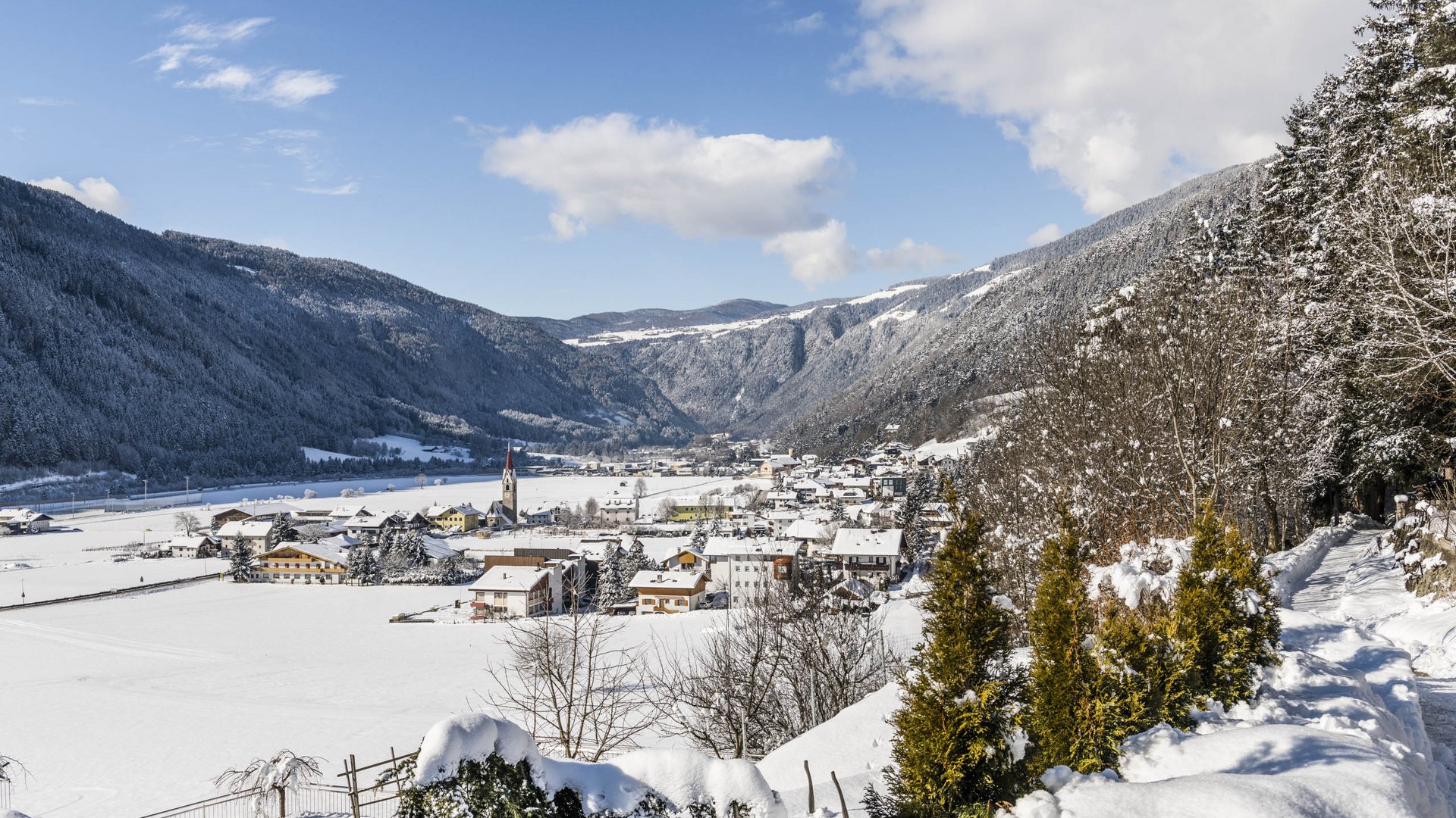 Vandoies: il cuore artigianale della Val Pusteria L'immagine mostra un villaggio innevato in una vasta valle, circondato da colline e montagne innevate. In primo piano c'è un sentiero coperto di neve con degli alberi, e sullo sfondo si vede una chiesa con una torre rossa che si erge tra le case del villaggio.