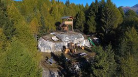 La zona ricreativa bunker di Spinga L'immagine mostra un'altura rocciosa in un'area boschiva, sulla quale è stata costruita una struttura in legno simile a un punto panoramico o a una piattaforma. In primo piano ci sono posti a sedere, attrezzature da gioco e una torre con scivolo, circondati da alberi verdi e folti e un cielo limpido e blu sullo sfondo.