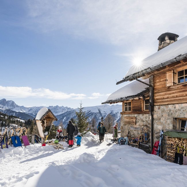 Kreuzwiesenalm Un rifugio di montagna si trova in un paesaggio invernale con montagne innevate sullo sfondo. Davanti al rifugio ci sono numerose slitte e diverse persone.