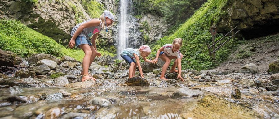 Le località dell’area vacanze Rio Pusteria L'immagine mostra tre bambini che giocano a piedi nudi in un ruscello poco profondo. Sullo sfondo si trova una grande cascata che scende da una parete rocciosa ripida, circondata da una rigogliosa foresta verde.