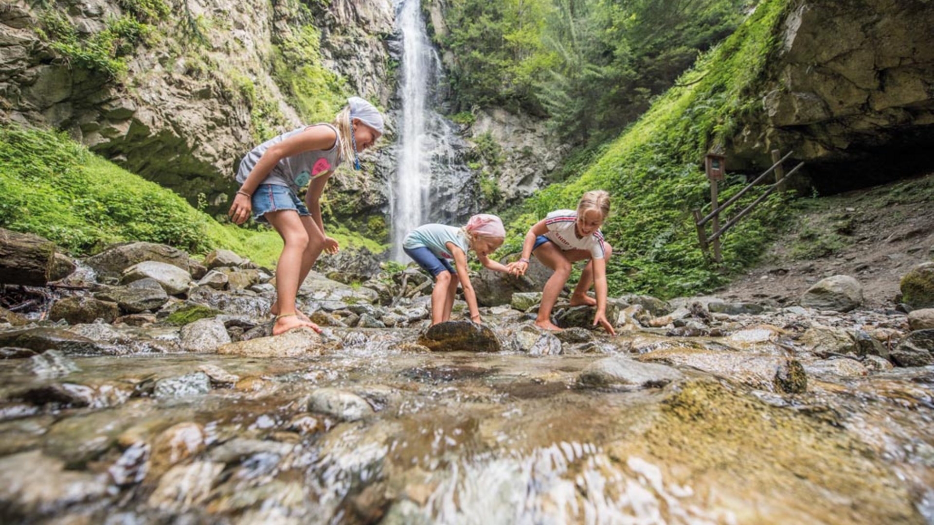 Il Sentiero degli Elfi a Vandoies di Sopra L'immagine mostra tre bambini che giocano a piedi nudi in un ruscello poco profondo. Sullo sfondo si trova una grande cascata che scende da una parete rocciosa ripida, circondata da una rigogliosa foresta verde.