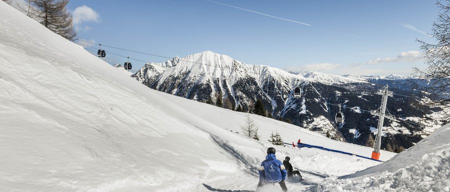Le località dell’area vacanze Rio Pusteria Persone che slittano su pista innevata con funivia e montagne sullo sfondo