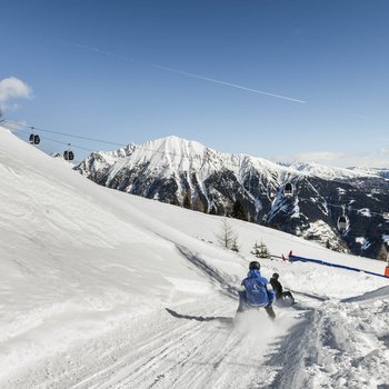 Benvenuti nell’Area Vacanze Sci e Malghe Rio Pusteria! Persone che slittano su pista innevata con funivia e montagne sullo sfondo