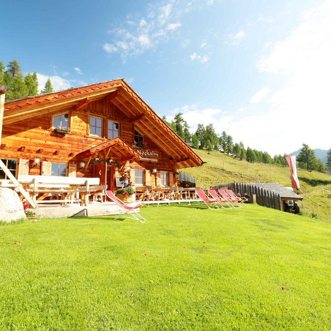 Nockalm L'immagine mostra una accogliente baita di legno chiamata "Nockalm" su un prato verde, circondata da sdraio e tavoli. Sullo sfondo si vedono colline boschive e un cielo limpido e azzurro.