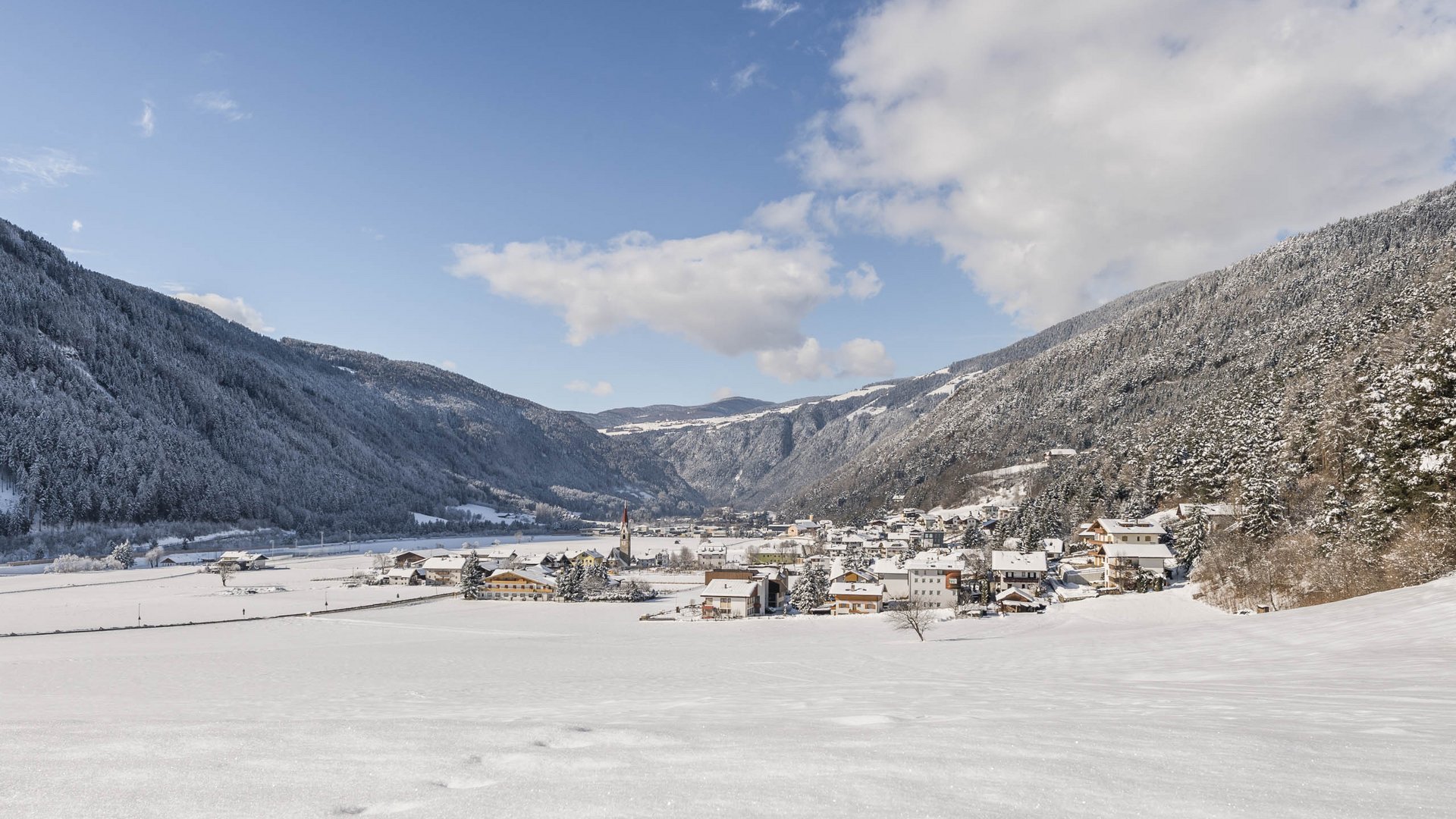 Vandoies: il cuore artigianale della Val Pusteria L'immagine mostra una valle innevata, in cui si trova un piccolo villaggio, circondato da montagne boschive coperte di neve. Il cielo è azzurro con alcune nuvole, e il paesaggio innevato appare calmo e pacifico.