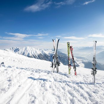 Benvenuti nell’Area Vacanze Sci e Malghe Rio Pusteria! Più sci piantati nella neve con montagne innevate e cielo blu sullo sfondo