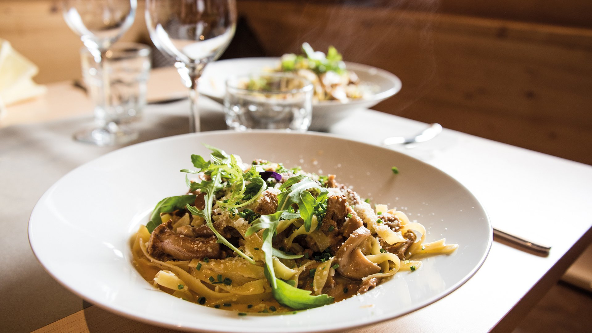 Alla scoperta delle malghe e dei rifugi di Maranza, Valles & Co. Nell'immagine c'è un piatto finemente preparato di pasta, guarnito con funghi ed erbe verdi fresche, servito su un piatto bianco in un ristorante accogliente con calici di vino e altri piatti sullo sfondo.