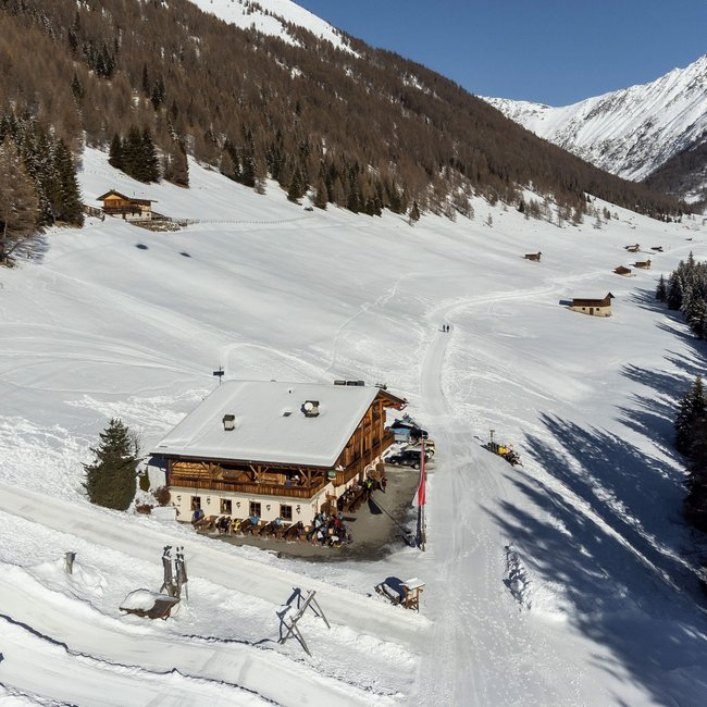 Großberghütte Un rifugio di montagna si trova in una valle innevata, circondato da pendii boscosi e da altre baite sparse. In primo piano si vedono delle persone davanti al rifugio, mentre un sentiero attraversa il paesaggio innevato.