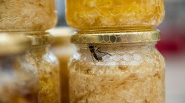 Shopping a Rio di Pusteria (BZ) L'immagine mostra un barattolo con favi di miele, sul cui coperchio è seduta un'ape. Il barattolo ha un'etichetta dorata con motivo a celle, e sullo sfondo si vedono altri barattoli di miele.