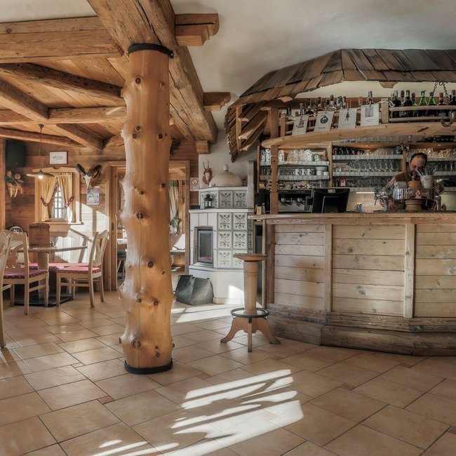 Großberghütte Una locanda rustica con travi di legno e tavoli con sedie imbottite. Sullo sfondo c'è un bancone con un cameriere e numerosi bicchieri e bottiglie sugli scaffali.