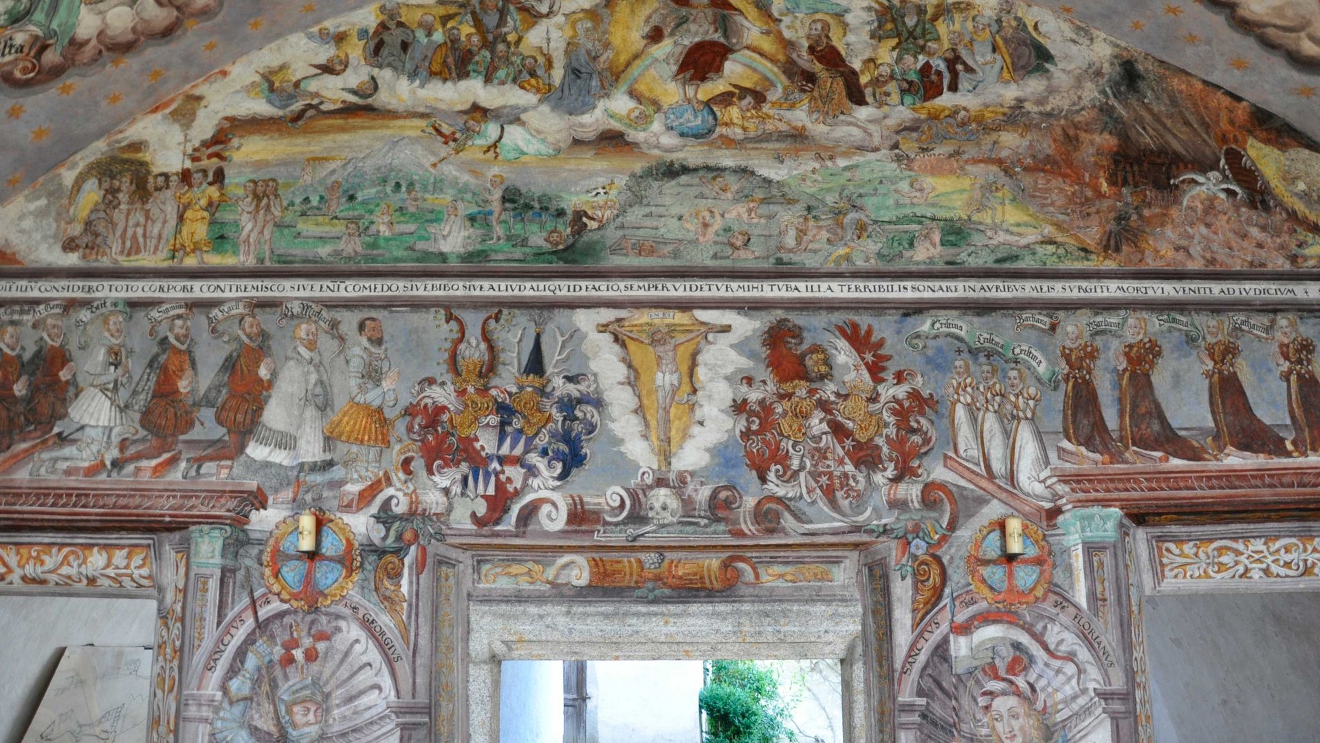 Alla scoperta di Castel Rodengo L'immagine mostra un affresco murale dettagliato in una stanza storica, che raffigura figure religiose e nobili, oltre a stemmi araldici. Le scene sono colorate e riccamente decorate.