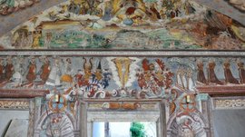 Alla scoperta di Castel Rodengo L'immagine mostra un affresco murale dettagliato in una stanza storica, che raffigura figure religiose e nobili, oltre a stemmi araldici. Le scene sono colorate e riccamente decorate.
