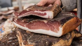 Shopping a Rio di Pusteria (BZ) L'immagine mostra una mano che tiene un grosso pezzo di pancetta. La pancetta ha una crosta scura speziata e mostra chiaramente gli strati bianchi di grasso e la carne rosso scuro all'interno. Diversi pezzi di pancetta sono impilati uno sopra l'altro su una base.