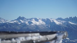 Valles - Jochtal - Steinermandl L'immagine mostra un vasto paesaggio montano con cime innevate sotto un cielo azzurro e limpido. In primo piano c'è una ringhiera di legno sfocata che dirige lo sguardo verso le maestose montagne sullo sfondo.