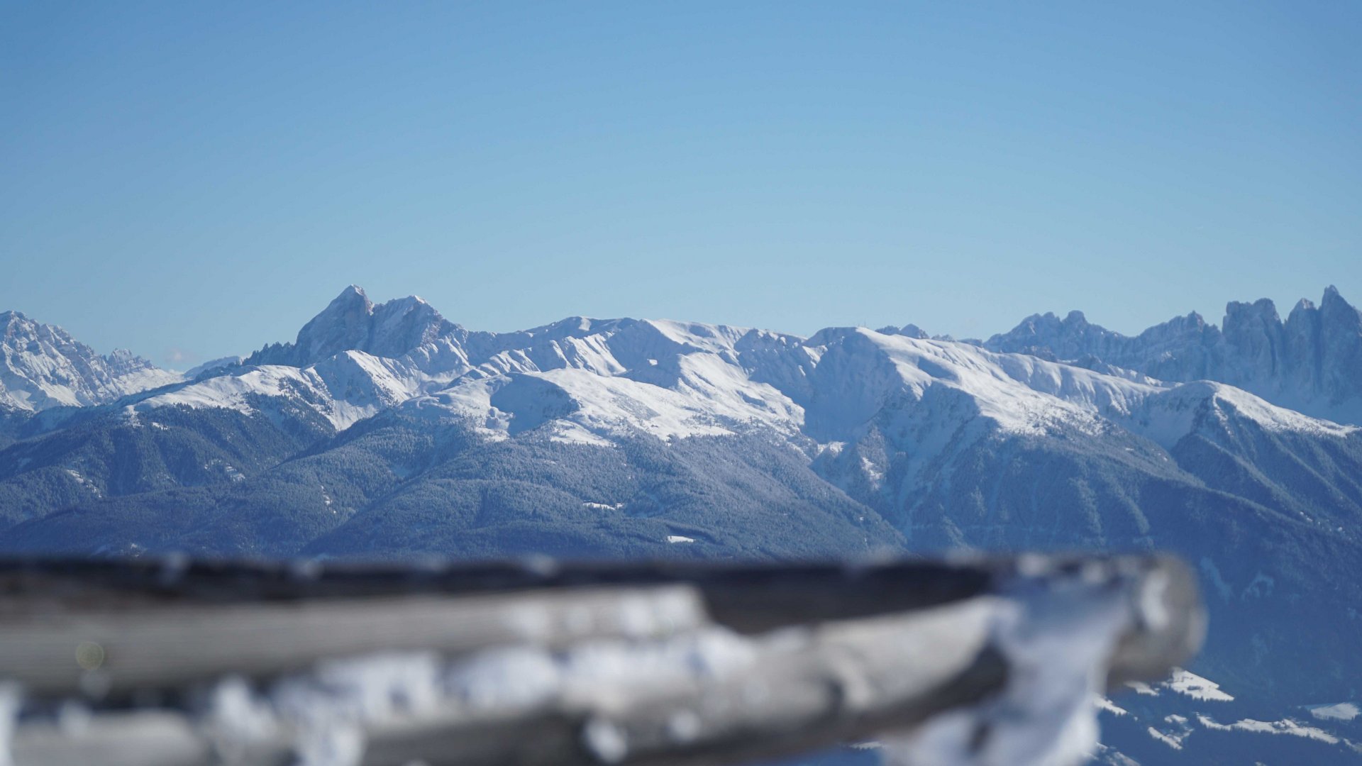 Valles - Jochtal - Steinermandl L'immagine mostra un vasto paesaggio montano con cime innevate sotto un cielo azzurro e limpido. In primo piano c'è una ringhiera di legno sfocata che dirige lo sguardo verso le maestose montagne sullo sfondo.