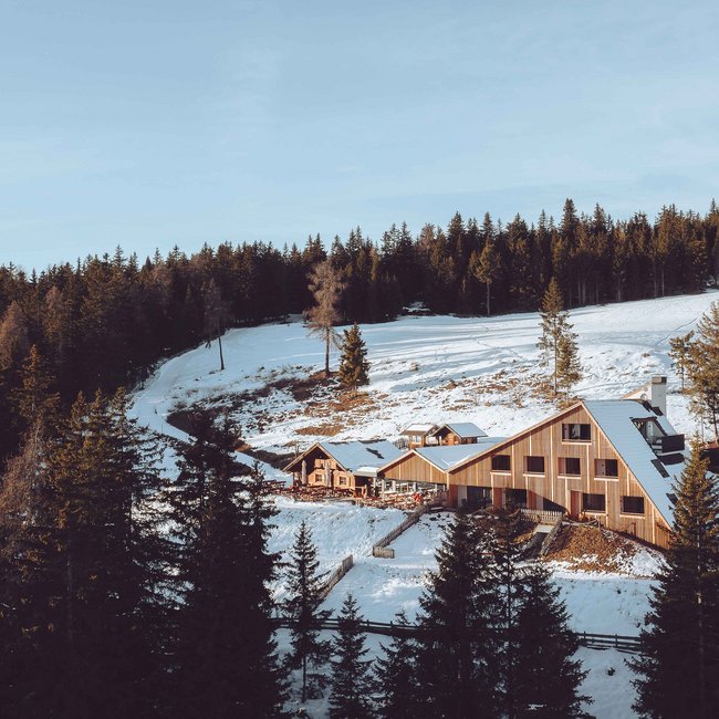 Oberhauserhütte L'immagine mostra un moderno rifugio di montagna in legno, circondato da colline innevate e da una fitta foresta. Il rifugio si trova in un pittoresco paesaggio invernale con abeti in primo piano e un cielo azzurro limpido sopra.