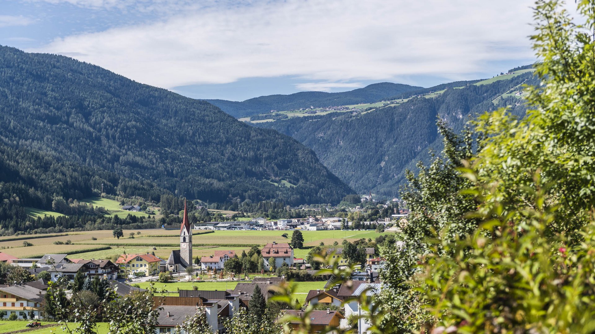 Vandoies: il cuore artigianale della Val Pusteria L'immagine mostra una valle con un piccolo villaggio, circondato da colline boschive. Al centro del villaggio si erge un campanile con tetto rosso, mentre sullo sfondo si estendono campi verdi e foreste fino alle colline.