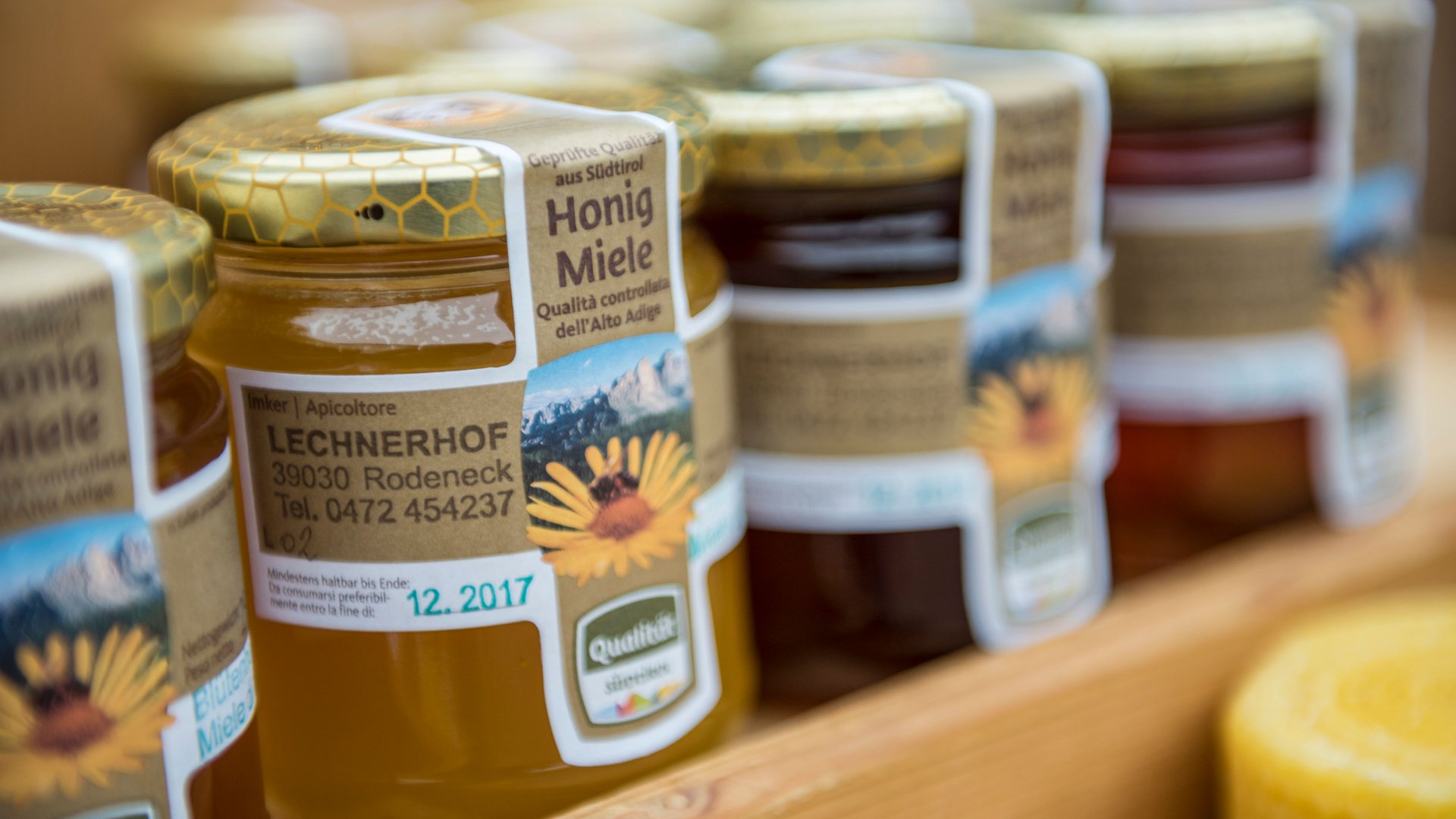 Shopping a Rio di Pusteria (BZ) L'immagine mostra diversi barattoli di miele disposti su una mensola. Sulle etichette dei barattoli è scritto "Honig Miele" insieme all'indirizzo e ai contatti di un apicoltore che lavora in una fattoria chiamata "Lechnerhof" a Rodengo in Alto Adige. I barattoli hanno coperchi dorati con un motivo a nido d'ape.