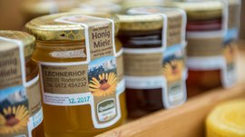 Shopping a Rio di Pusteria (BZ) L'immagine mostra diversi barattoli di miele disposti su una mensola. Sulle etichette dei barattoli è scritto "Honig Miele" insieme all'indirizzo e ai contatti di un apicoltore che lavora in una fattoria chiamata "Lechnerhof" a Rodengo in Alto Adige. I barattoli hanno coperchi dorati con un motivo a nido d'ape.