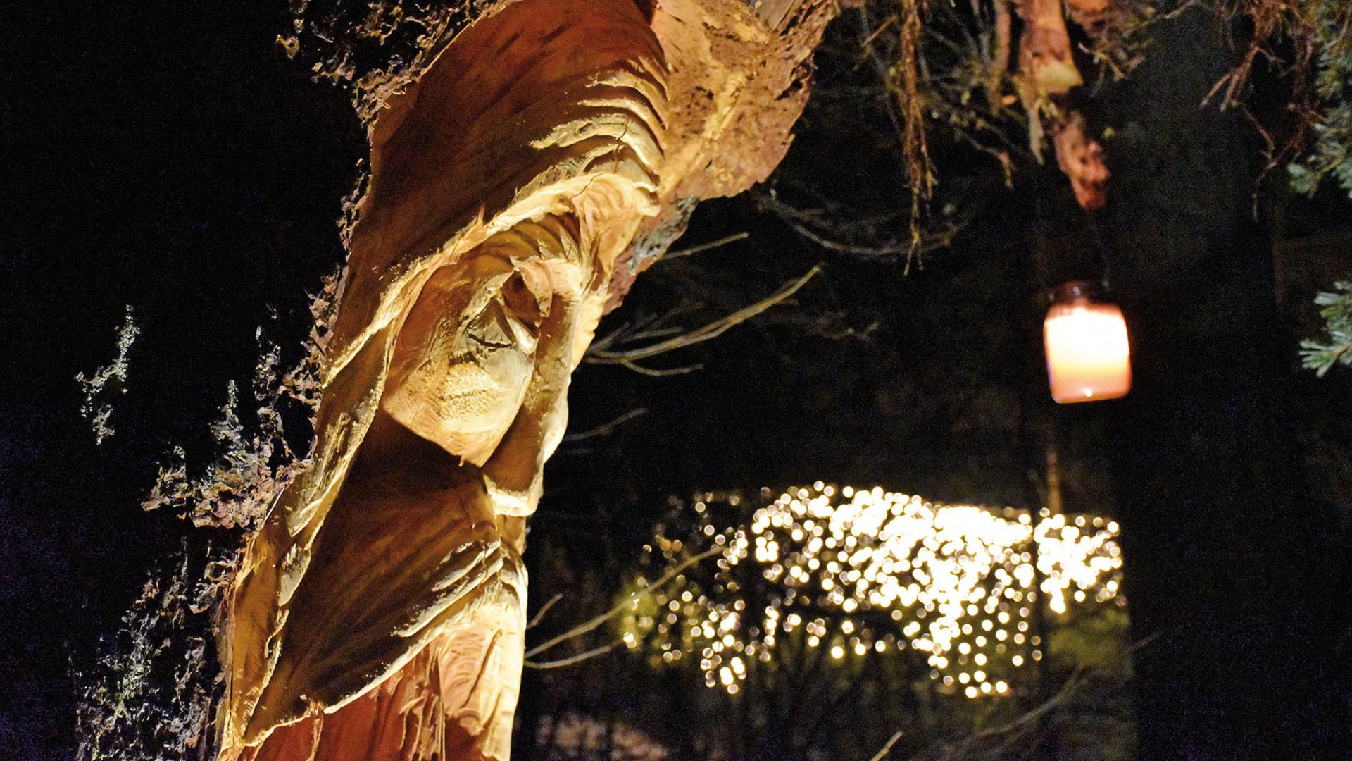 L’Avvento di Terento L'immagine mostra una figura intagliata nel legno, integrata in un tronco d'albero, presumibilmente una rappresentazione religiosa come Maria. La scena è illuminata in modo suggestivo e sullo sfondo si vedono catene di luci sfocate e una candela accesa in un bicchiere, che creano un'atmosfera calda e festosa.