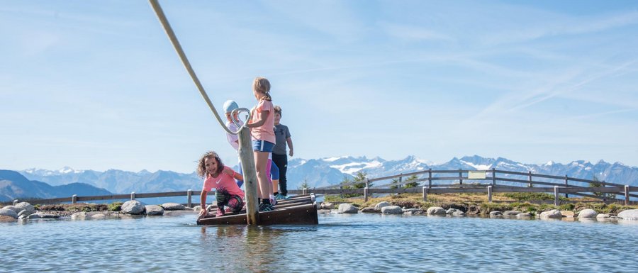 Le località dell’area vacanze Rio Pusteria L'immagine mostra diversi bambini che stanno su una piccola zattera, tirata con una corda su uno stagno poco profondo. Sullo sfondo si vedono delle montagne che completano il cielo azzurro limpido e il paesaggio pittoresco.