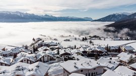 Conoscete già Maranza? L'immagine mostra un villaggio con case innevate su una collina, circondato da una copertura di nebbia. Sullo sfondo si vedono imponenti cime montuose innevate sotto un cielo parzialmente nuvoloso.