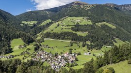 Quando si dice idilliaco… ecco a voi Fundres L'immagine mostra un paesaggio verde e collinare con un piccolo villaggio situato in una valle. L'ambiente è circondato da fitte foreste e montagne, mentre il cielo è limpido e blu, con qualche nuvola bianca sparsa.