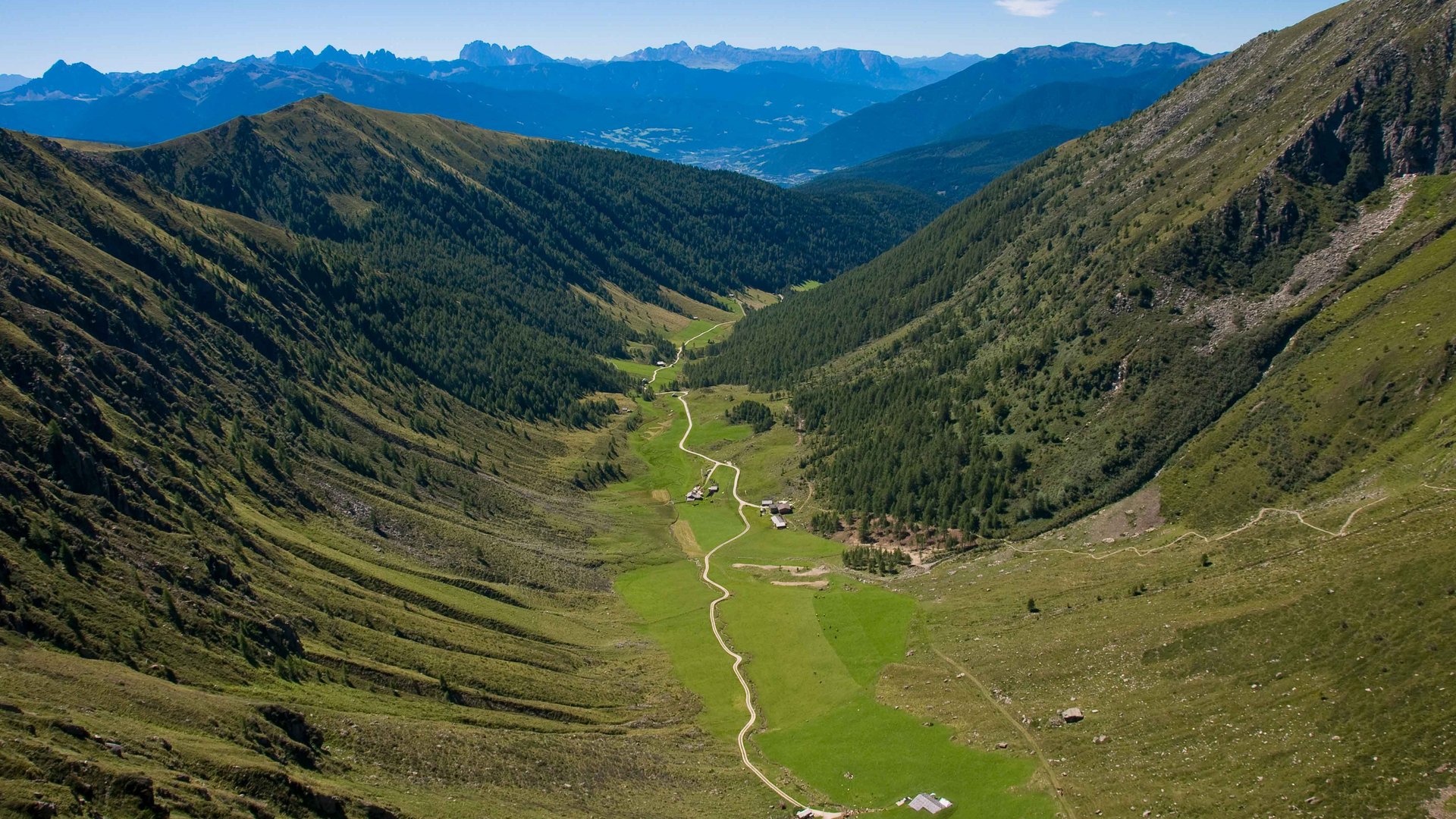 La Valle di Altafossa, presso Maranza L'immagine mostra una valle ampia che si estende tra due pendii montuosi alti e boscosi. Un sentiero stretto e tortuoso attraversa la valle verde, passando accanto ad alcune baite sparse. Sullo sfondo si possono vedere le maestose vette delle Alpi che dominano l'orizzonte.