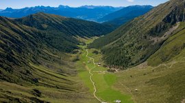 La Valle di Altafossa, presso Maranza L'immagine mostra una valle ampia che si estende tra due pendii montuosi alti e boscosi. Un sentiero stretto e tortuoso attraversa la valle verde, passando accanto ad alcune baite sparse. Sullo sfondo si possono vedere le maestose vette delle Alpi che dominano l'orizzonte.