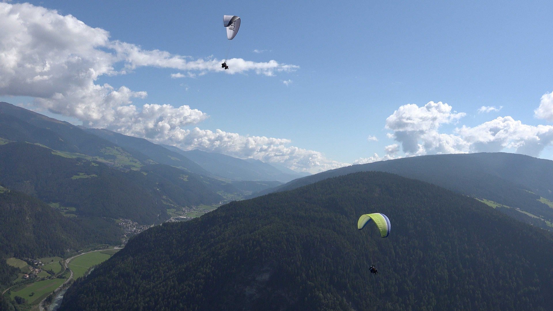 Parapendio in Alto Adige L'immagine mostra due parapendii che volano sopra un vasto paesaggio montano con colline densamente boschive. Il cielo è limpido con alcune nuvole sparse e sullo sfondo si estendono valli verdi e altre montagne.