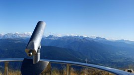 La piattaforma panoramica Gitschberga a Maranza (BZ) L'immagine mostra una vista panoramica da una vetta di montagna con un cannocchiale al centro e una croce di vetta a destra. Sullo sfondo si estendono ampie valli verdi e montagne sotto un cielo chiaro e azzurro.