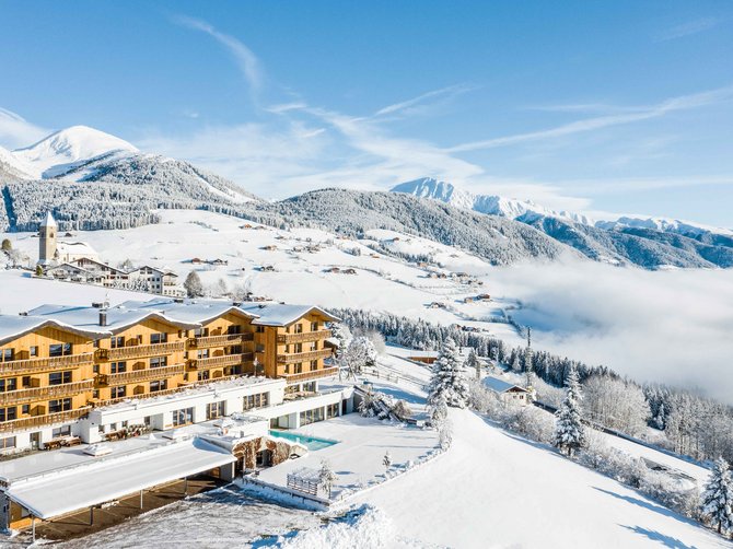 Benvenuti nell’Area Vacanze Sci e Malghe Rio Pusteria! L'immagine mostra un grande hotel in un paesaggio invernale coperto di neve. Sullo sfondo si vedono montagne innevate, foreste e una piccola chiesa, mentre l'hotel è illuminato da un sole splendente.