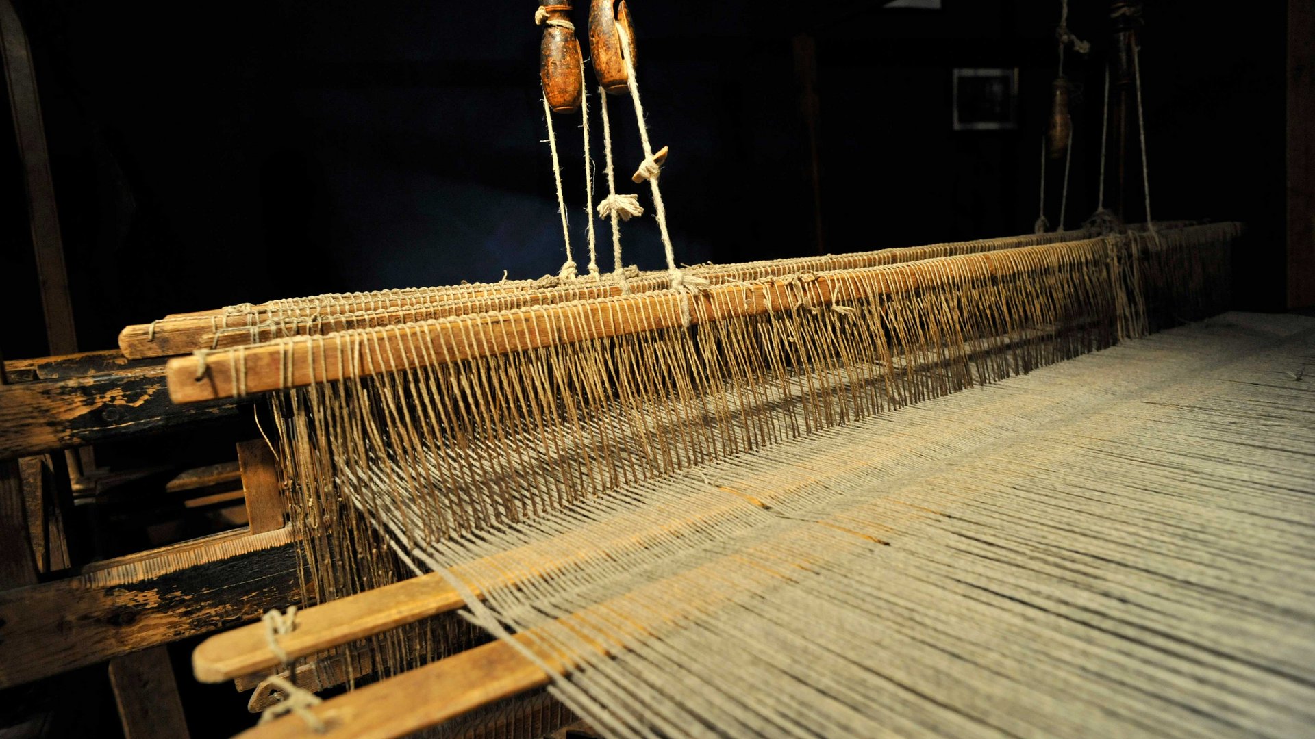 Il Mondo del Loden a Vandoies L'immagine mostra un vecchio telaio in legno con fili tesi strettamente uno accanto all'altro. Il telaio è esposto in un ambiente buio, il che rende ben visibili i dettagli della meccanica e dei fili.