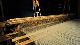 Il Mondo del Loden a Vandoies L'immagine mostra un vecchio telaio in legno con fili tesi strettamente uno accanto all'altro. Il telaio è esposto in un ambiente buio, il che rende ben visibili i dettagli della meccanica e dei fili.