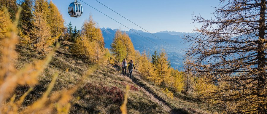 Le località dell’area vacanze Rio Pusteria L'immagine mostra una funivia che attraversa un sentiero utilizzato dagli escursionisti. Il paesaggio è autunnale, con alberi dorati, e sullo sfondo si vedono montagne sotto un cielo azzurro limpido.