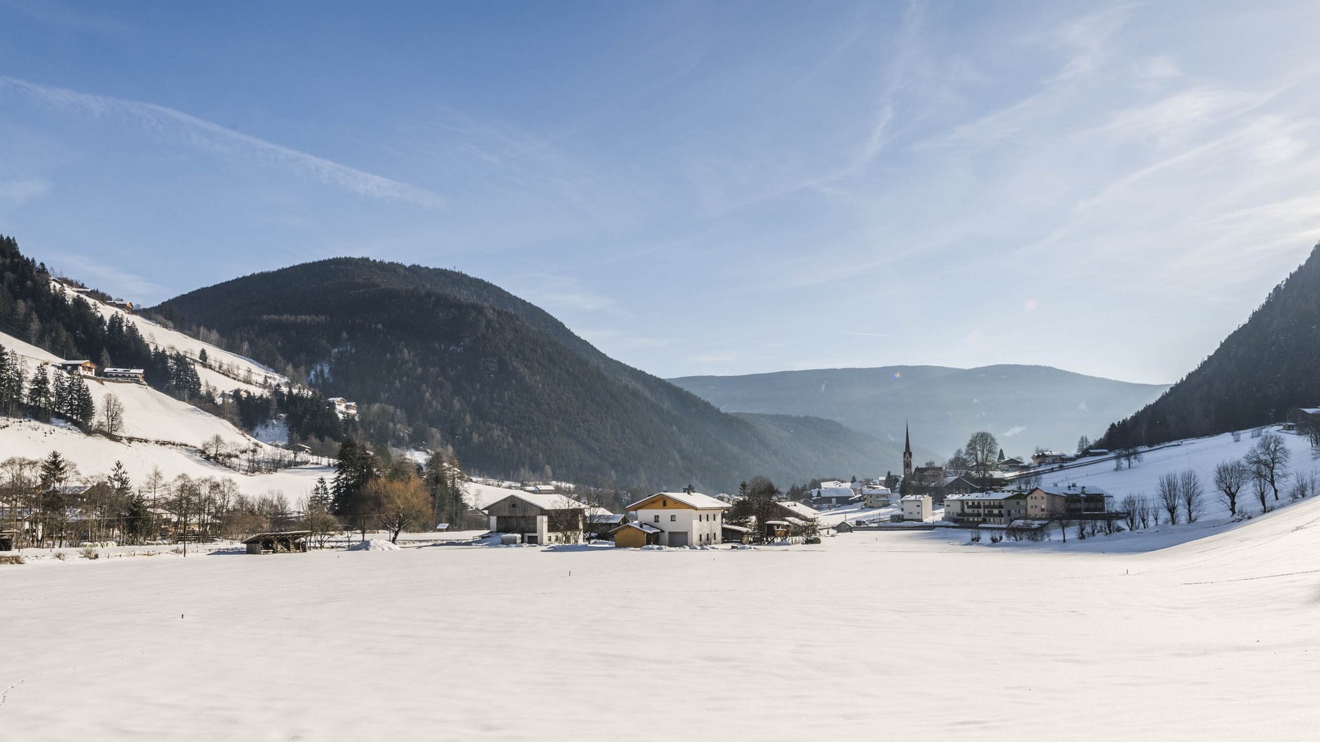 Quando si dice idilliaco… ecco a voi Fundres L'immagine mostra una valle innevata con un piccolo villaggio al centro, circondato da colline e boschi coperti di neve. Il cielo è limpido e blu, e il sole splende luminoso, proiettando lunghe ombre sulla neve.