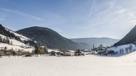 Quando si dice idilliaco… ecco a voi Fundres L'immagine mostra una valle innevata con un piccolo villaggio al centro, circondato da colline e boschi coperti di neve. Il cielo è limpido e blu, e il sole splende luminoso, proiettando lunghe ombre sulla neve.
