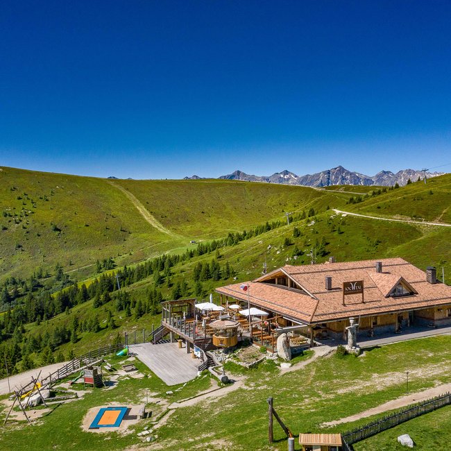 Bergrestaurant Jochtal L'immagine mostra un grande rifugio di montagna con un'ampia terrazza, circondato da dolci colline verdi e sentieri escursionistici in un paesaggio montano rurale sotto un cielo azzurro e limpido.