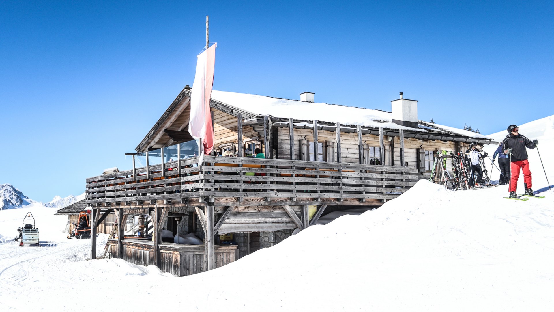 Rifugi e malghe di Rio Pusteria Nell'immagine si vede un rustico chalet da sci in legno, circondato dalla neve, sulla cui terrazza sono seduti alcuni ospiti; in primo piano c'è uno sciatore vicino a degli sci appoggiati.