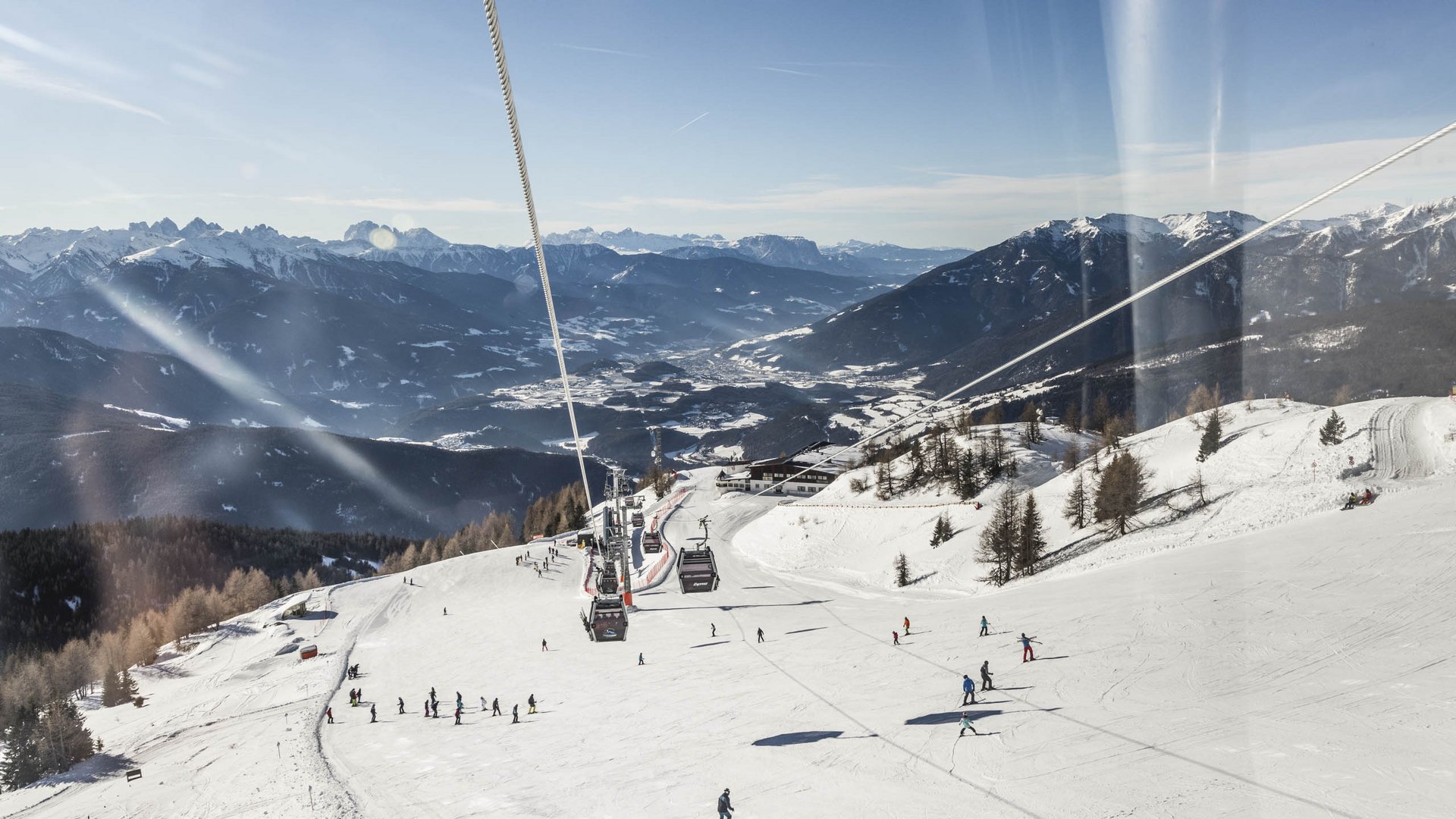 Sciare in Alto Adige: Rio Pusteria L'immagine mostra una vasta pista da sci con numerosi sciatori che scendono lungo il pendio. Sopra la pista corre una funivia con cabine, e sullo sfondo si estende un panorama impressionante di montagne e valli innevate sotto un cielo azzurro e limpido.