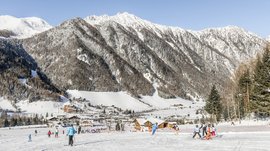 Valles & il villaggio alpino più bello dell’Alto Adige L'immagine mostra un paesaggio montano innevato con ripide pendici montane sullo sfondo e sciatori in primo piano. Diverse persone scendono la pista mentre in fondo alla valle si estende un piccolo villaggio con case di legno.