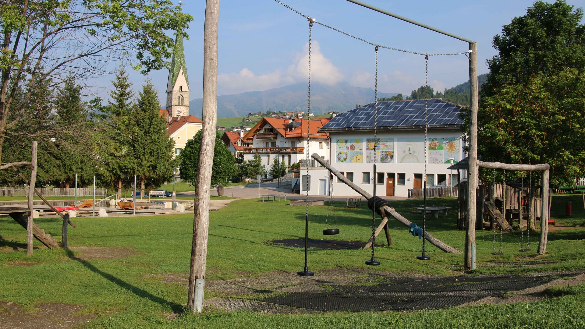 Il Parco Active a Terento L'immagine mostra un parco giochi con altalene in primo piano, situato su un prato verde. Sullo sfondo si vede un edificio con pannelli solari, pareti dipinte e una chiesa con una torre appuntita, circondati da alberi e un paesaggio collinare.
