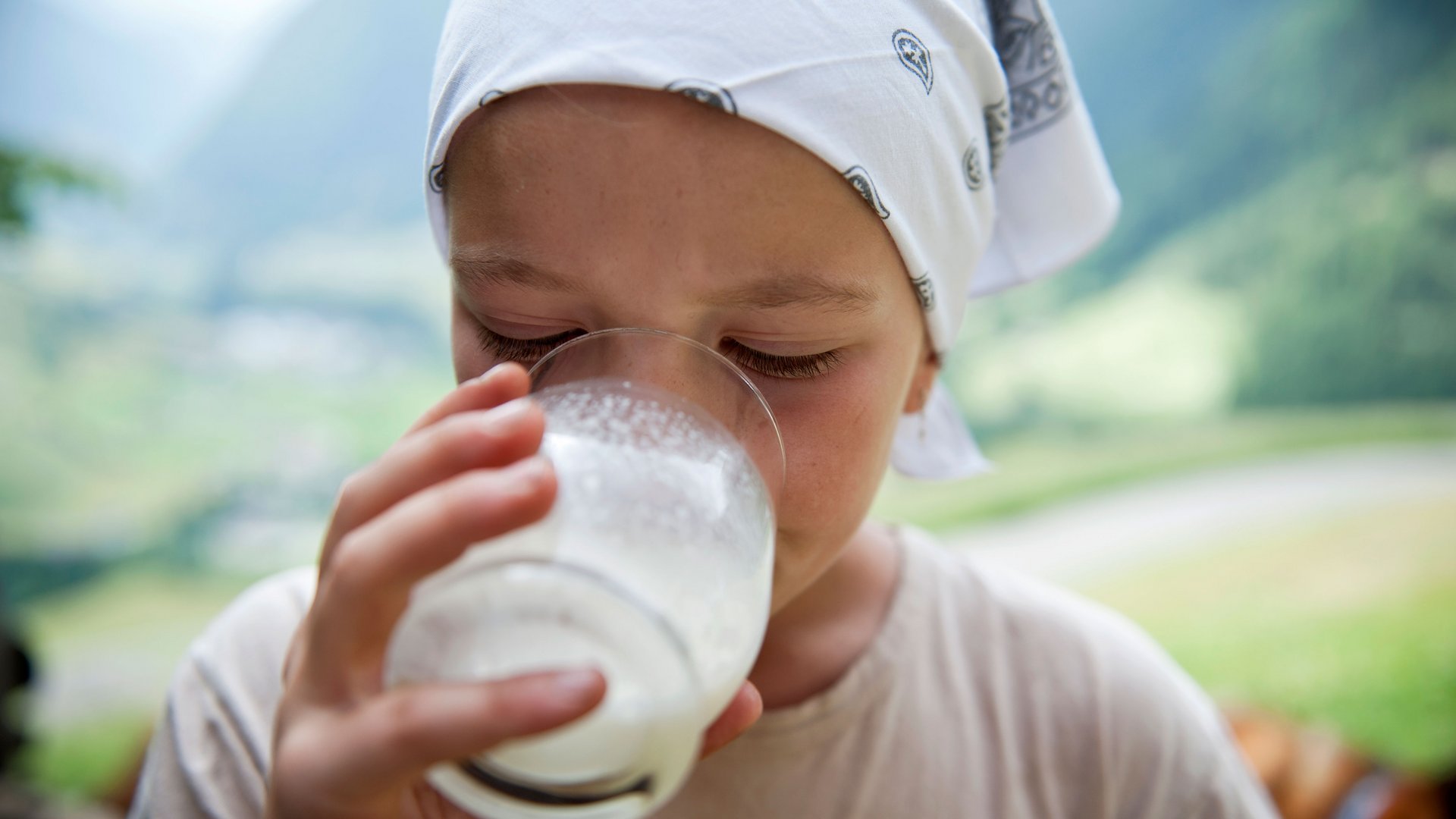 Il Sentiero dei masi di Fundres Nell'immagine si vede un bambino che beve da un bicchiere di latte. Il bambino indossa un foulard bianco con un motivo, e sullo sfondo si riconosce un paesaggio montano verde sfocato.